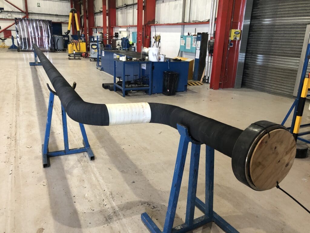 Long bent pipeline section with black Neoprene rubber coating and capped end, staged at LJF Custom Coatings workshop